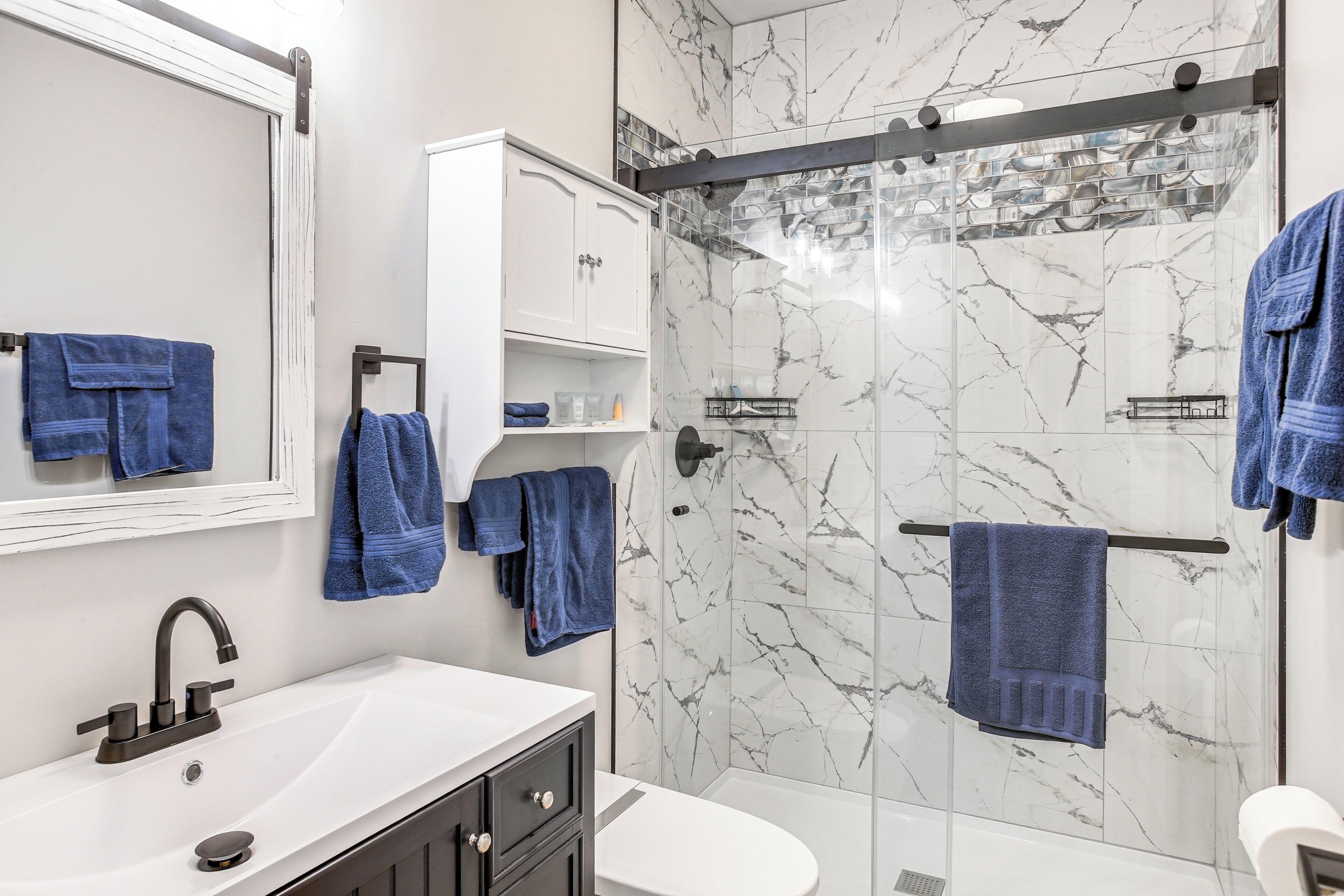 A modern bathroom with a white sink, dark cabinets, a glass shower enclosure, and marble walls.