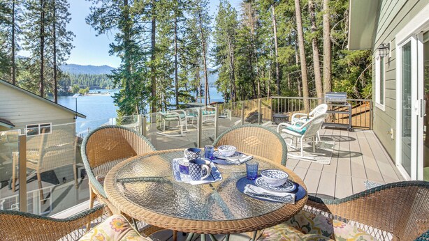 A deck with wicker chairs and a glass table set for dining, overlooking a lake and surrounded by tall trees. - Lake Lover's Paradise: Waterfront Home in Hayden! (Hayden)
