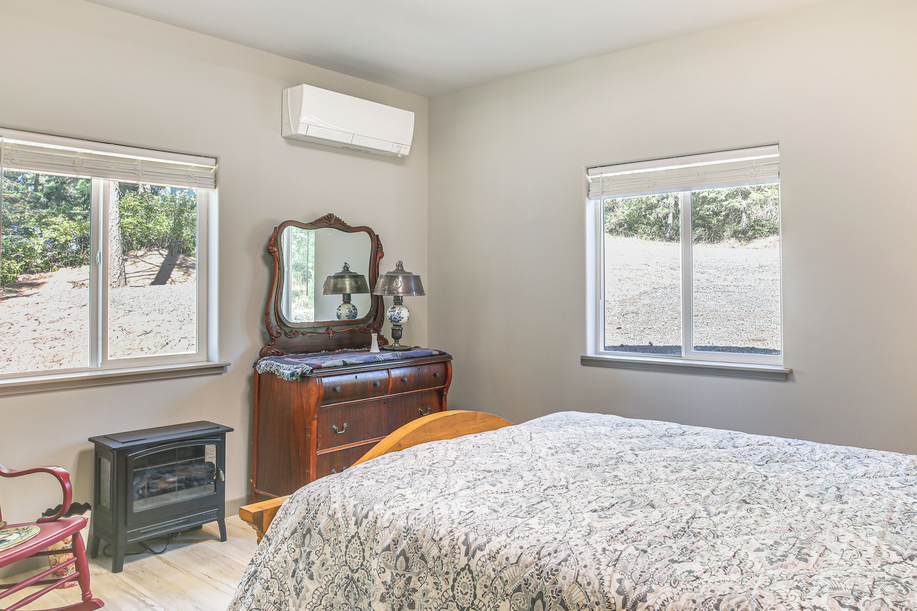 Une chambre à coucher comprenant un lit, une commode, un miroir et deux fenêtres donnant sur des arbres.
