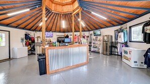 A spacious indoor area with a high wooden ceiling and a central wooden structure. There are various items and equipment on the walls and floor, including a refrigerator, a washing machine, and a counter with a display. - Private Dock & Slip: Kentucky Lake Cottage! (Buchanan)