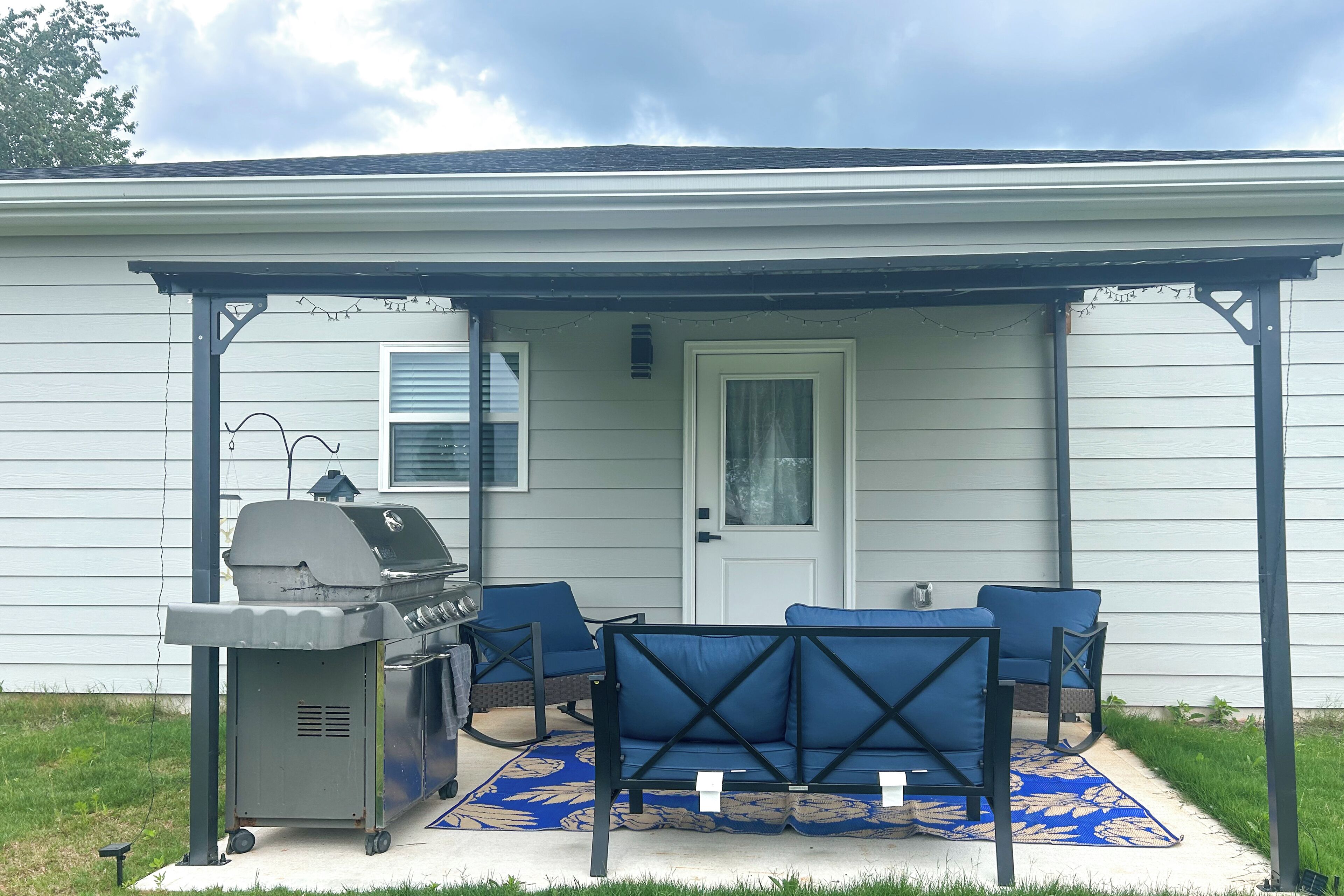 Eine überdachte Terrasse mit Grill, blauem Gartenmöbel und einem gemusterten Teppich.