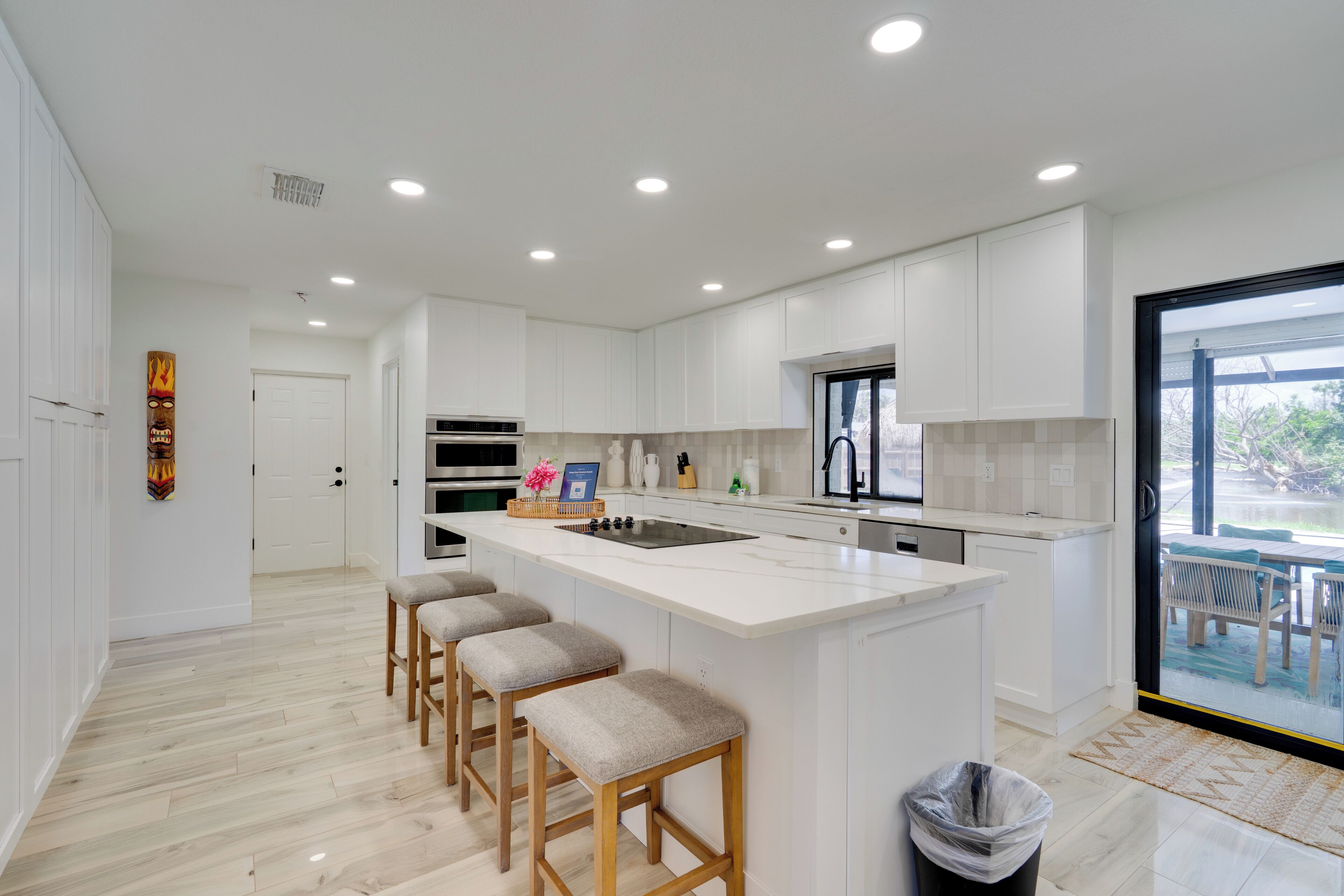 Una cocina moderna con armarios blancos, una isla central con taburetes y vistas a través de un ventanal.