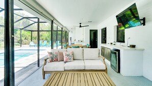 Amplia sala de estar con un ventanal grande, piso de madera, un sofá con cojines, televisor montado en la pared y una zona de cocina con lavavajillas.