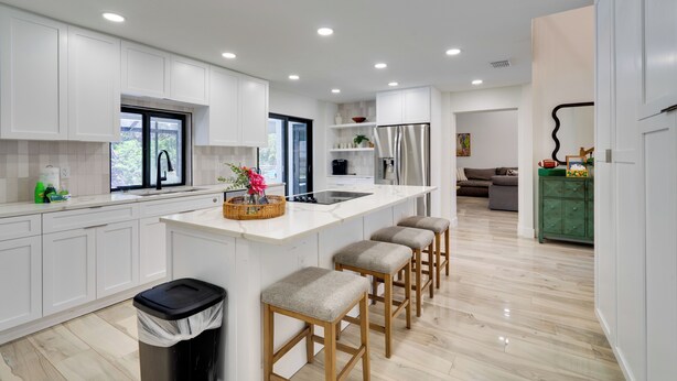 Una cocina moderna con armarios blancos, una isla central con taburetes, un refrigerador de acero inoxidable y un ventanal que proporciona luz natural.