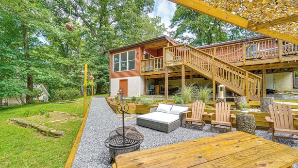 Una terraza de madera con mobiliario exterior, una fogata y un área cubierta con una escalera que conduce a un segundo nivel.
