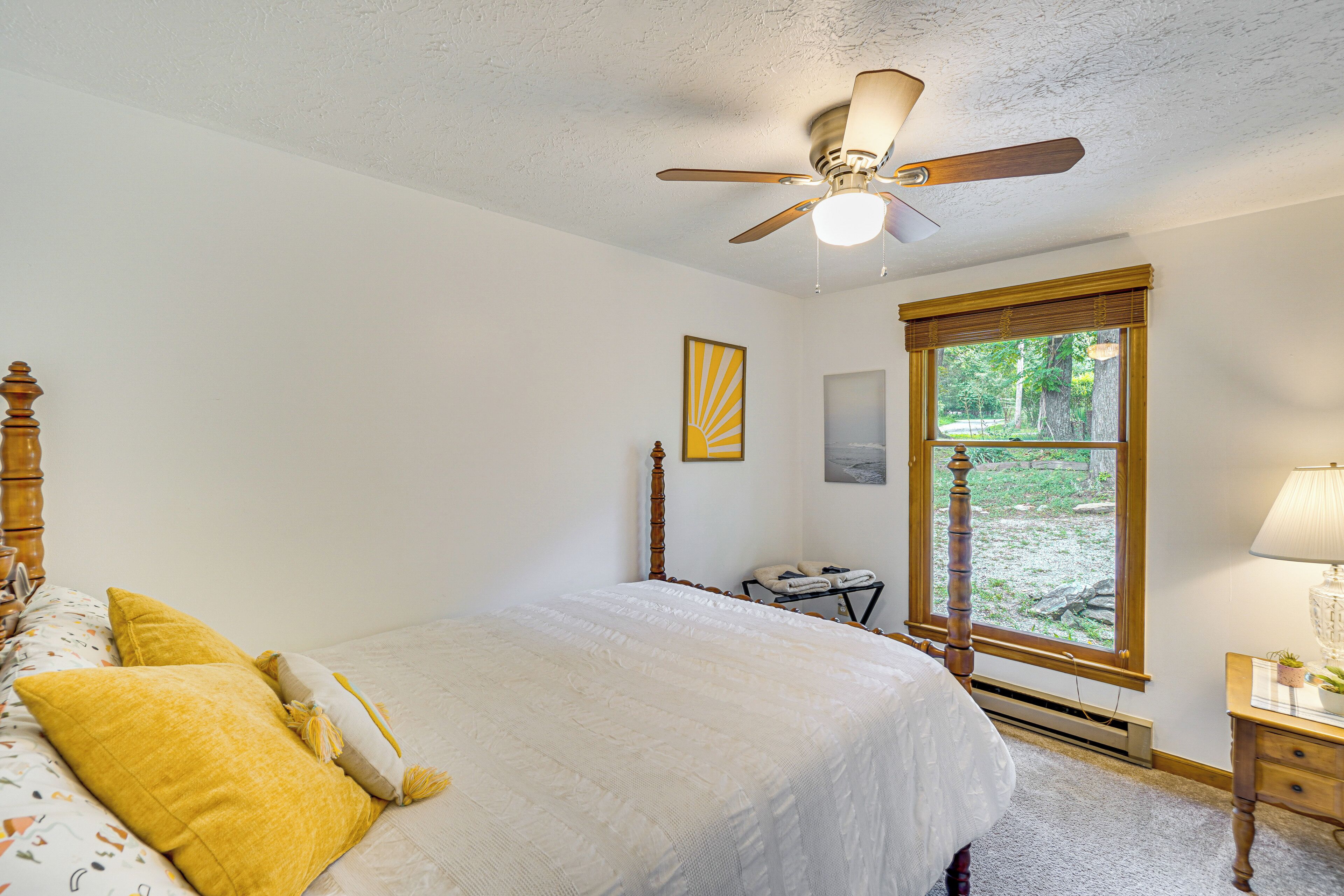 Ein Schlafzimmer mit Bett, Deckenventilator, Fenster mit Blick ins Grüne und Nachttisch mit Lampe.