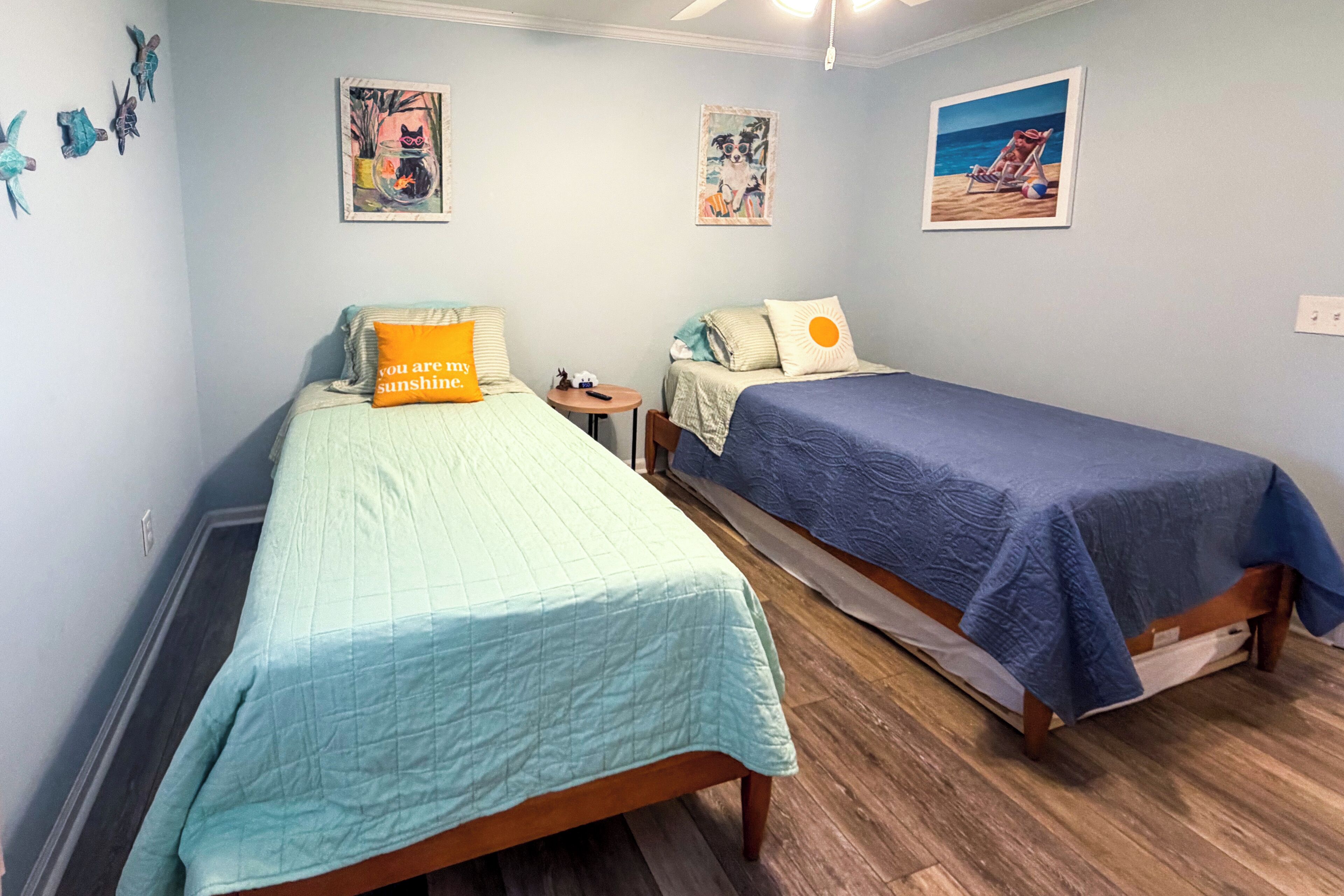 A room with two beds, each with a different colored bedspread, a small table, and a wall with three framed pictures.