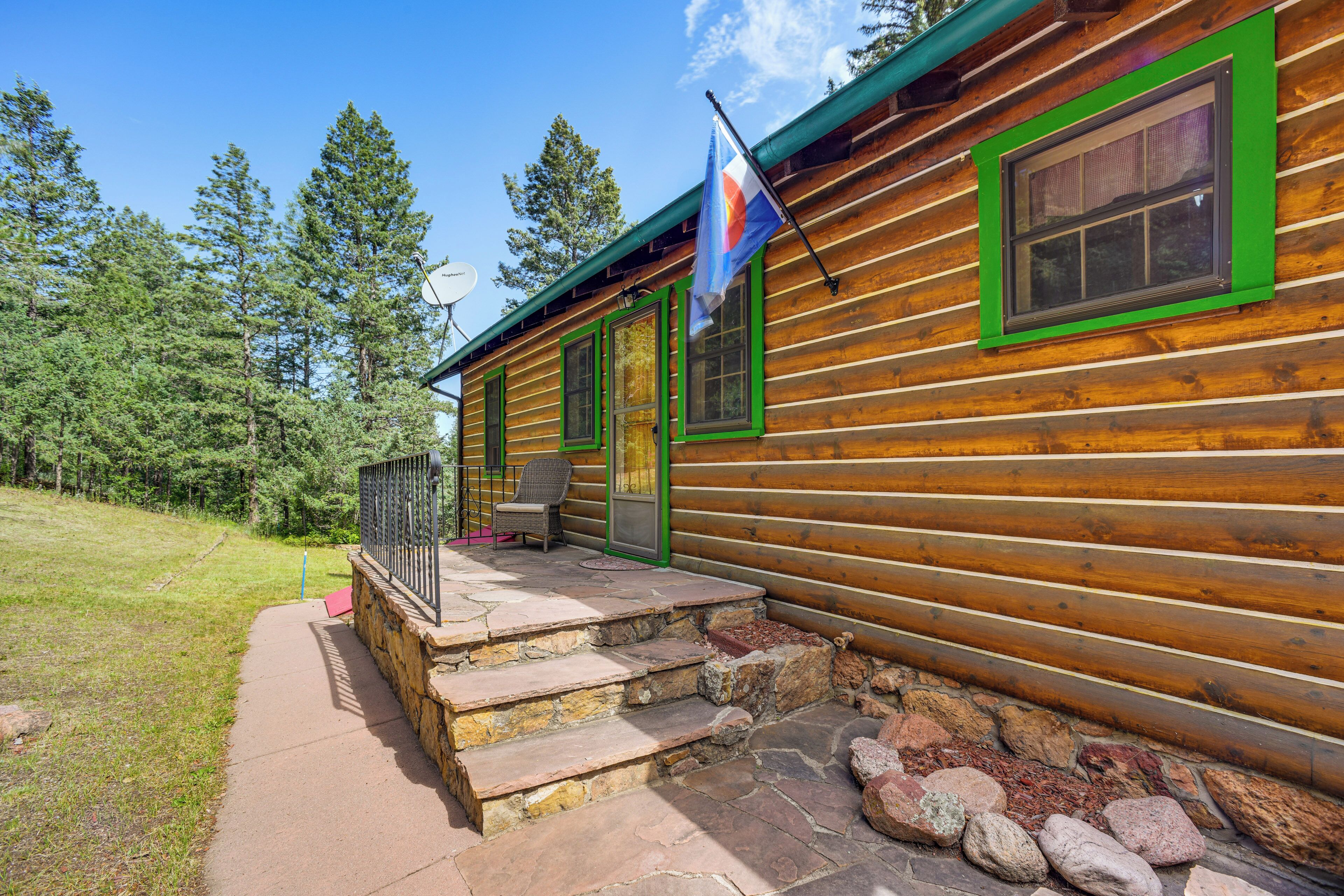 A log cabin with a stone staircase leading to a porch, a flag, and a view of the surrounding forest.
