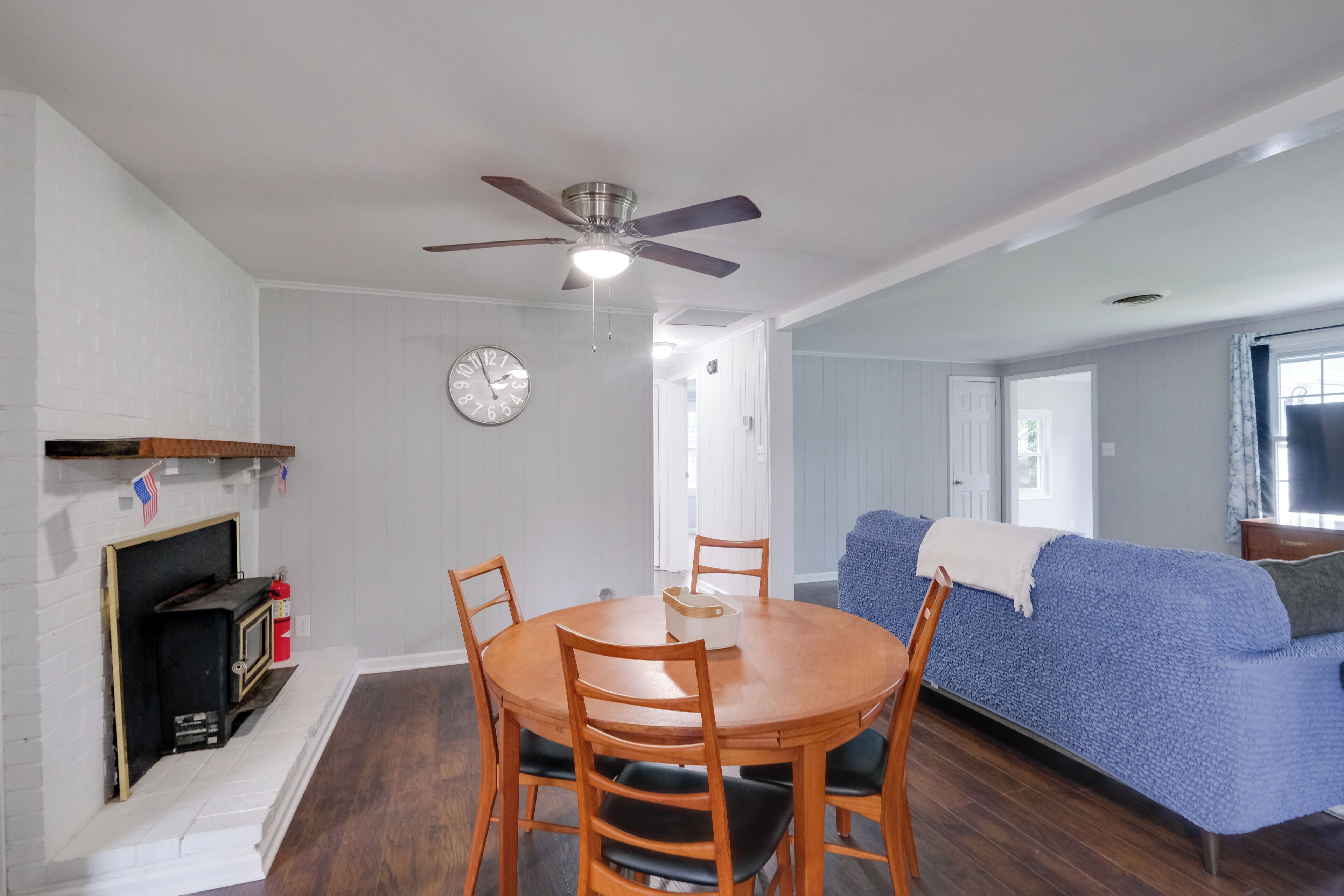 A dining area with a round wooden table and chairs, a ceiling fan, a clock on the wall, and a blue sofa in the background.