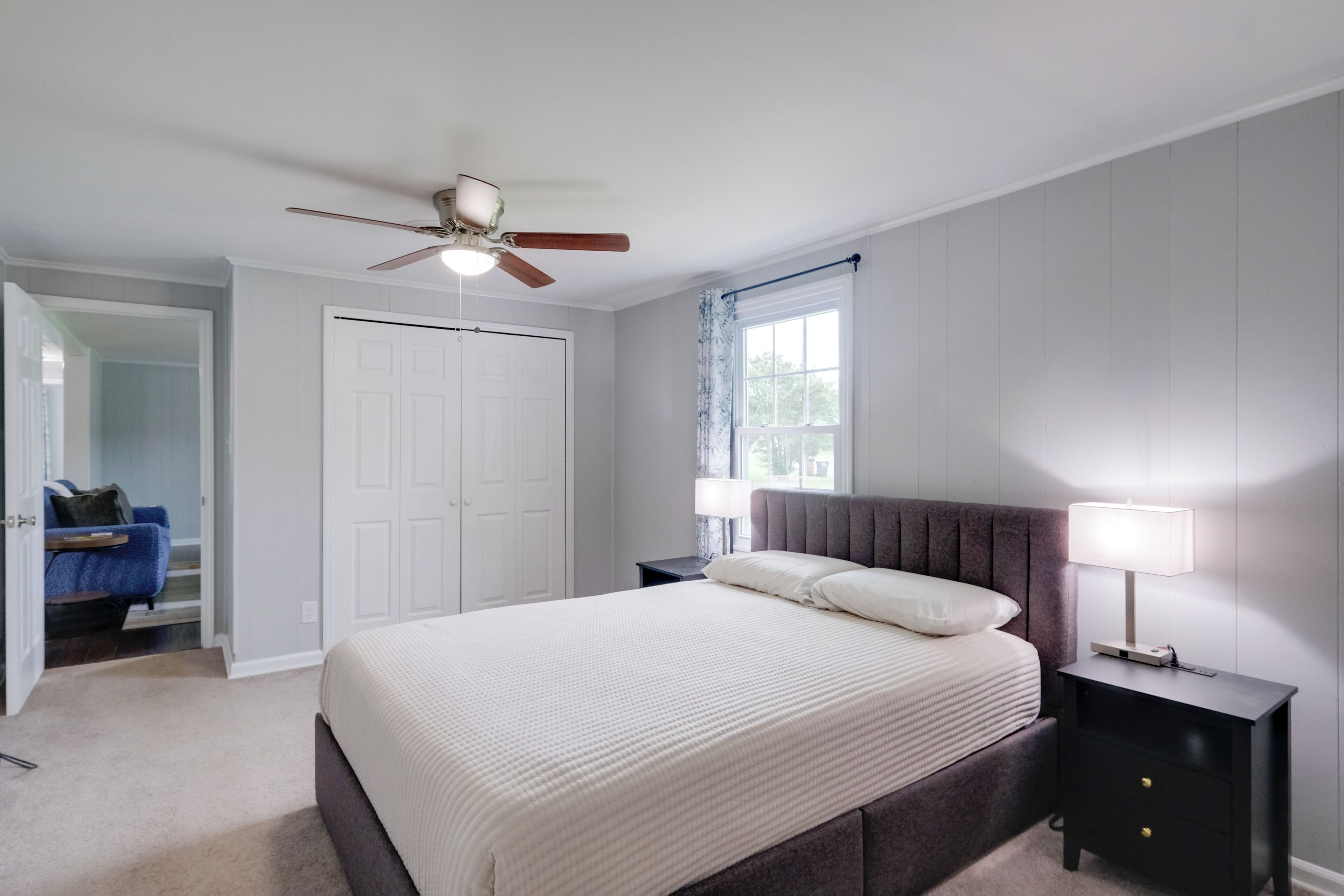A bedroom with a bed, a nightstand, a ceiling fan, a window with curtains, and a door leading to another room.