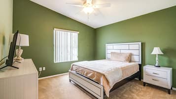 Quarto com cama, mesa de cabeceira, luminária, ventoinha no teto, janela com persianas e escrivaninha com luminária.