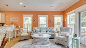 A living room with a round coffee table, two sofas, a dining area with white chairs, and large windows with white shutters.