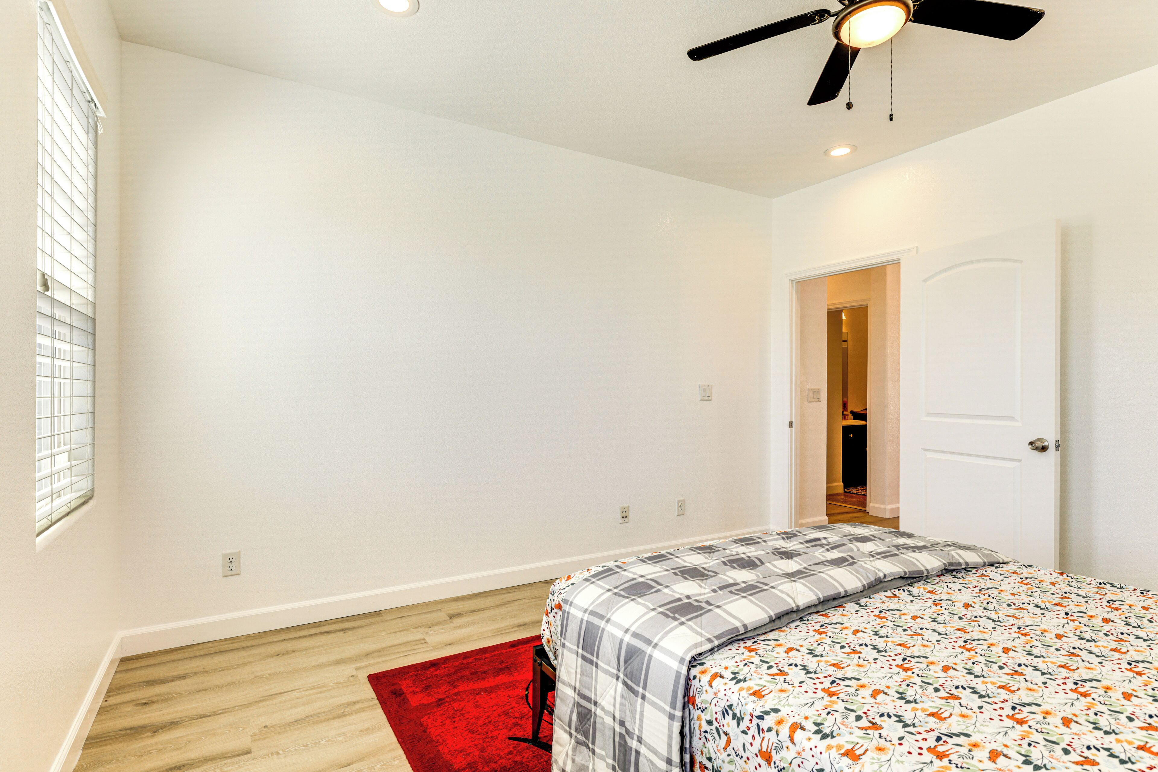 A bedroom with a bed, a red rug, a ceiling fan, and a door leading to another room.