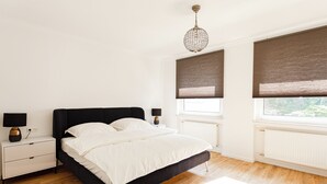 A modern bedroom with a black bed, white bedding, a nightstand with a lamp, and a large window with blinds.
