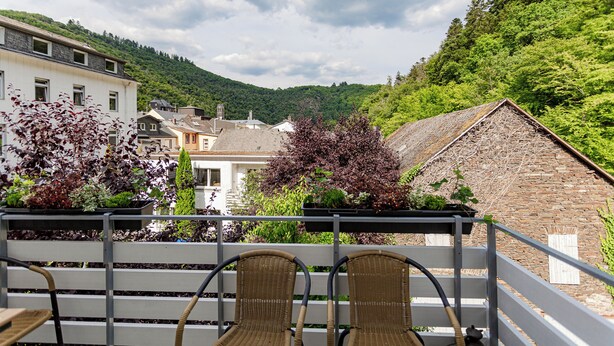 A balcony with wicker chairs overlooking a mountainous landscape, a stone building, and a variety of plants.