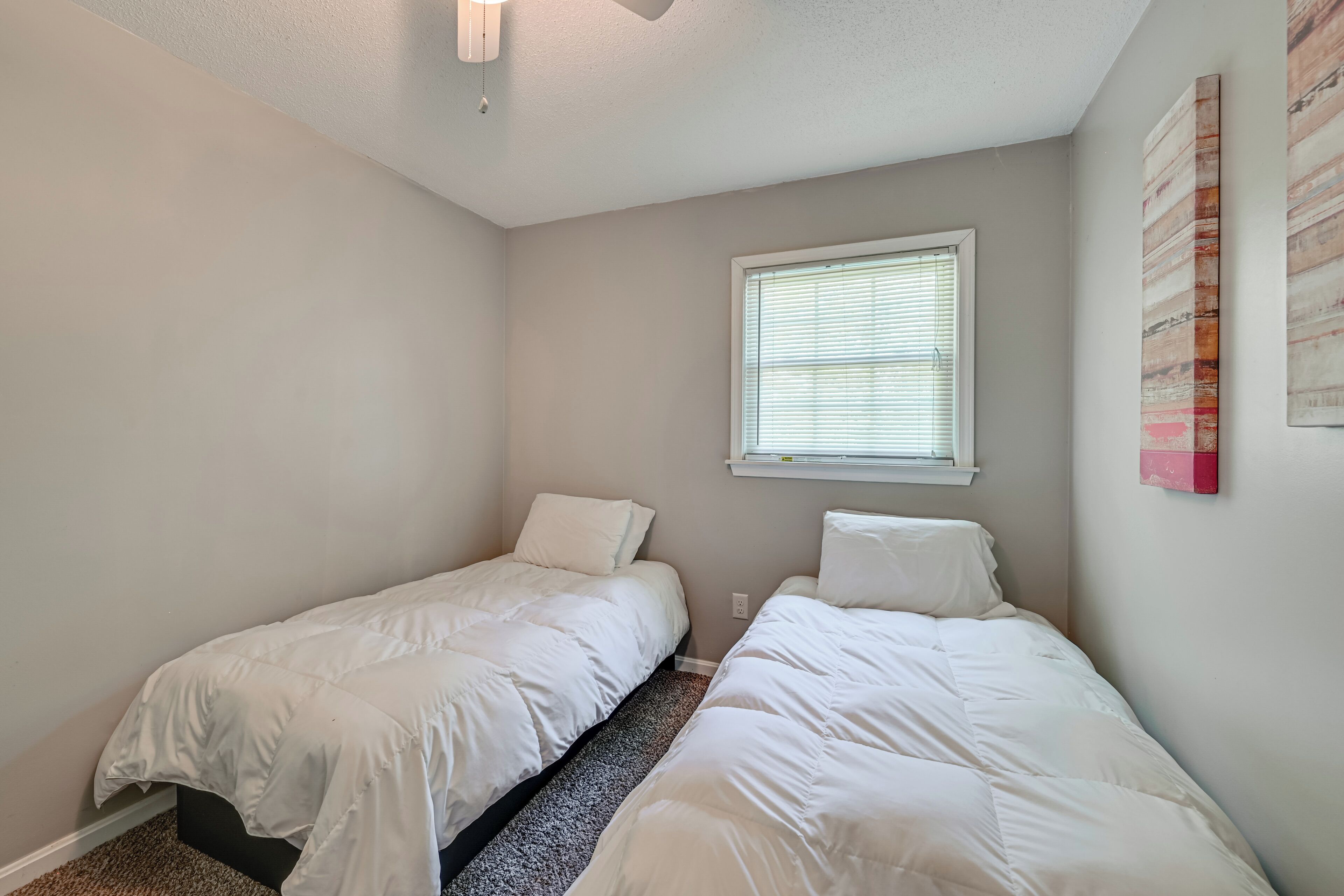 A small, clean room with two single beds, a window with blinds, and a wall-mounted artwork.