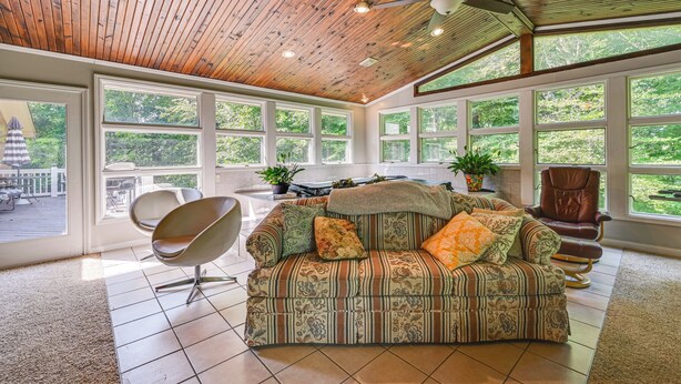 Een ruime woonkamer met houten plafond, grote ramen, een bank, een fauteuil en een eettafel.