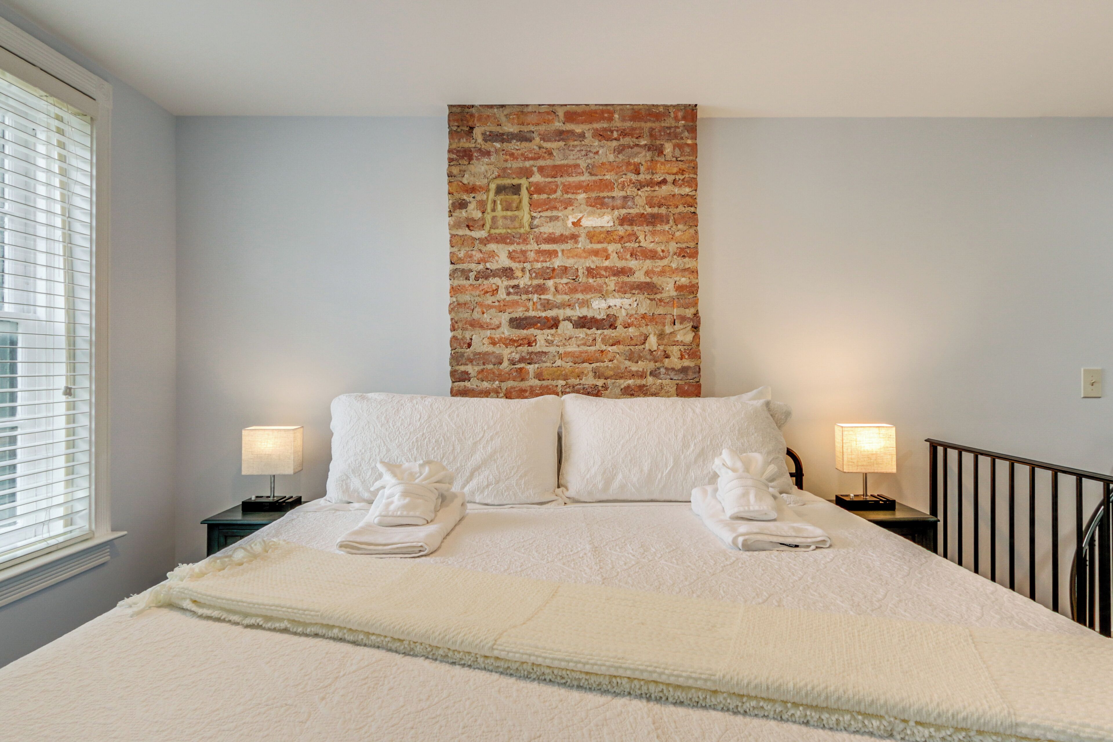 A neatly made bed with white linens, two bedside lamps, and a brick accent wall.