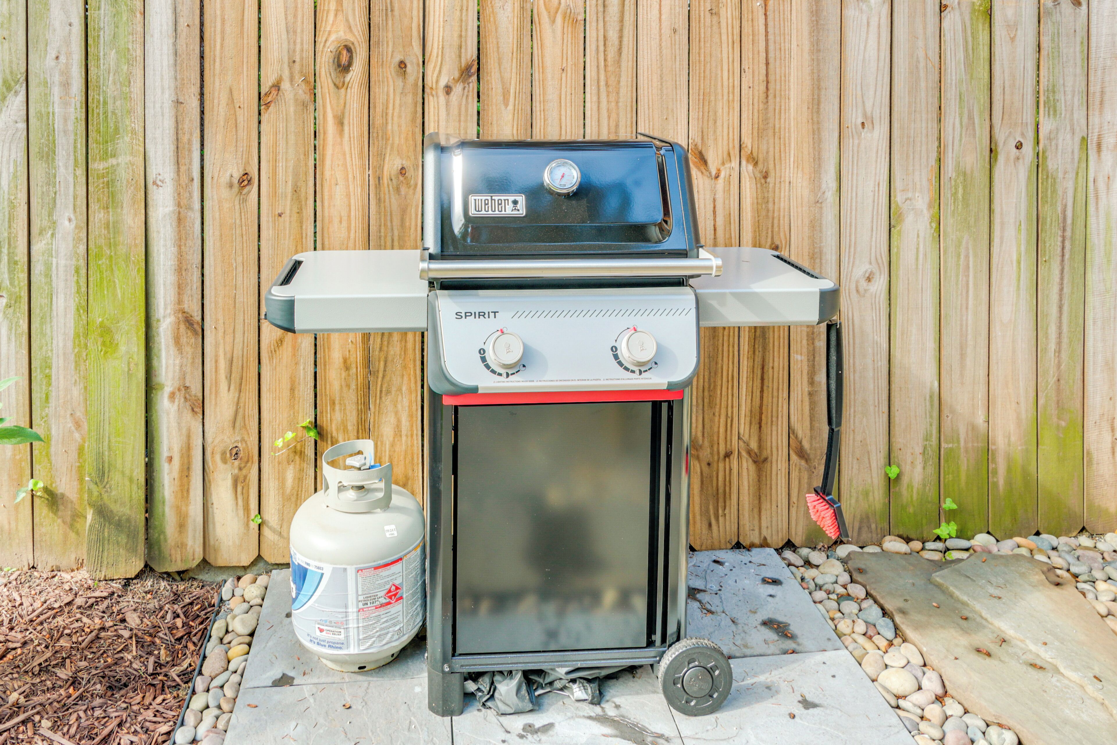 Una parrilla de gas portátil con horno integrado, un tanque de propano y una rueda para facilitar el transporte, frente a una valla de madera.