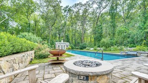 A stone patio with a fire pit, outdoor seating, and a swimming pool surrounded by lush greenery. - Pool, Gardens & Hot Tub: Luxe Sandy Hook Home! (Sandy Hook)