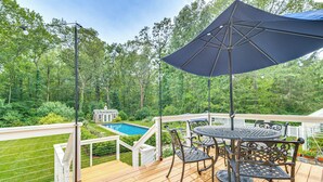 A wooden deck with a table and chairs, a large umbrella, and a view of a swimming pool and greenery. - Pool, Gardens & Hot Tub: Luxe Sandy Hook Home! (Sandy Hook)