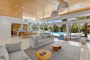 Uma sala de estar moderna e integrada com sofá, mesa de centro e mesa de jantar, grandes janelas com vista para a piscina, e uma cozinha ao fundo.