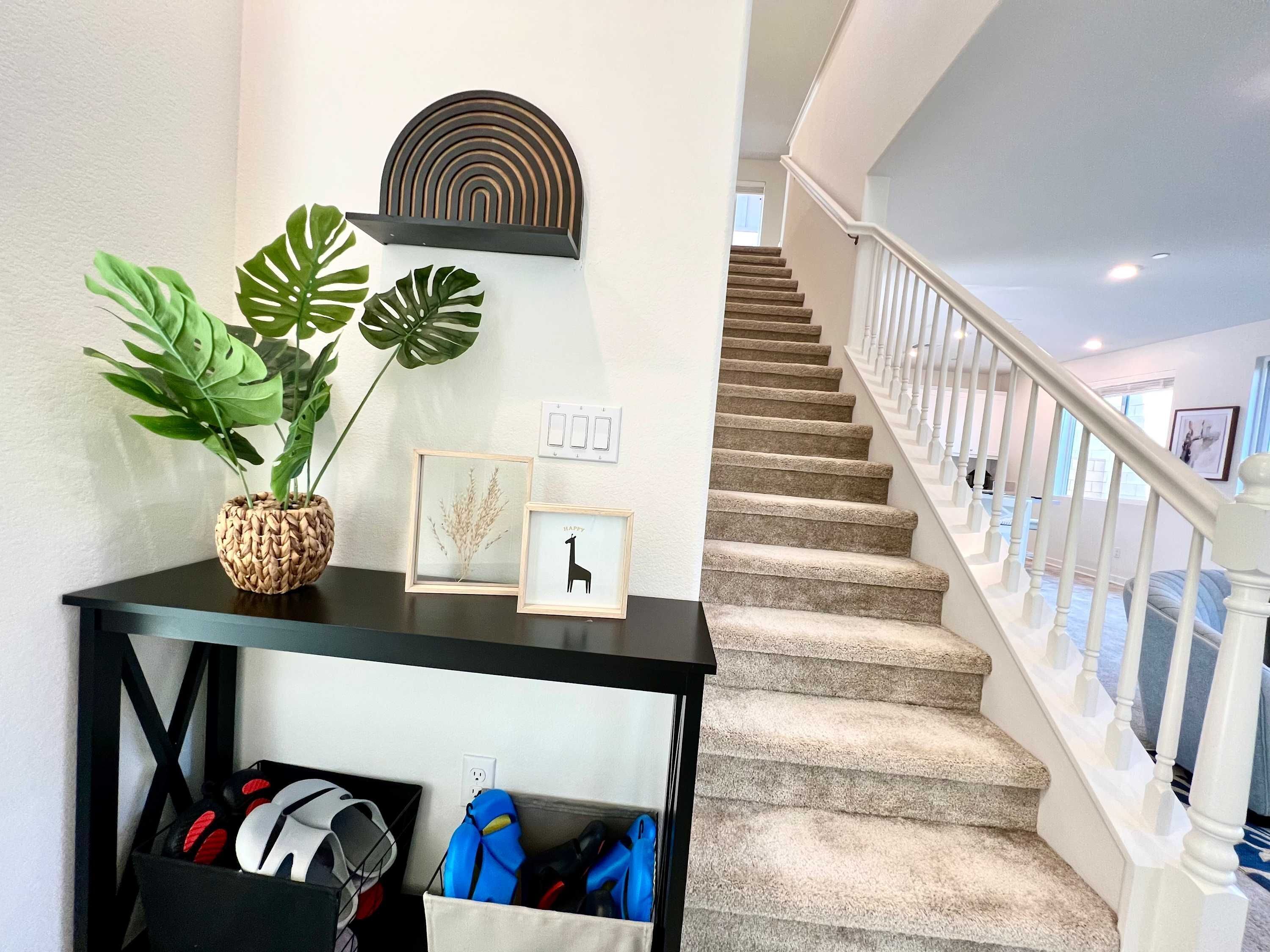 Una escalera con barandilla blanca, una consola negra con plantas y cuadros, y un montón de zapatos.