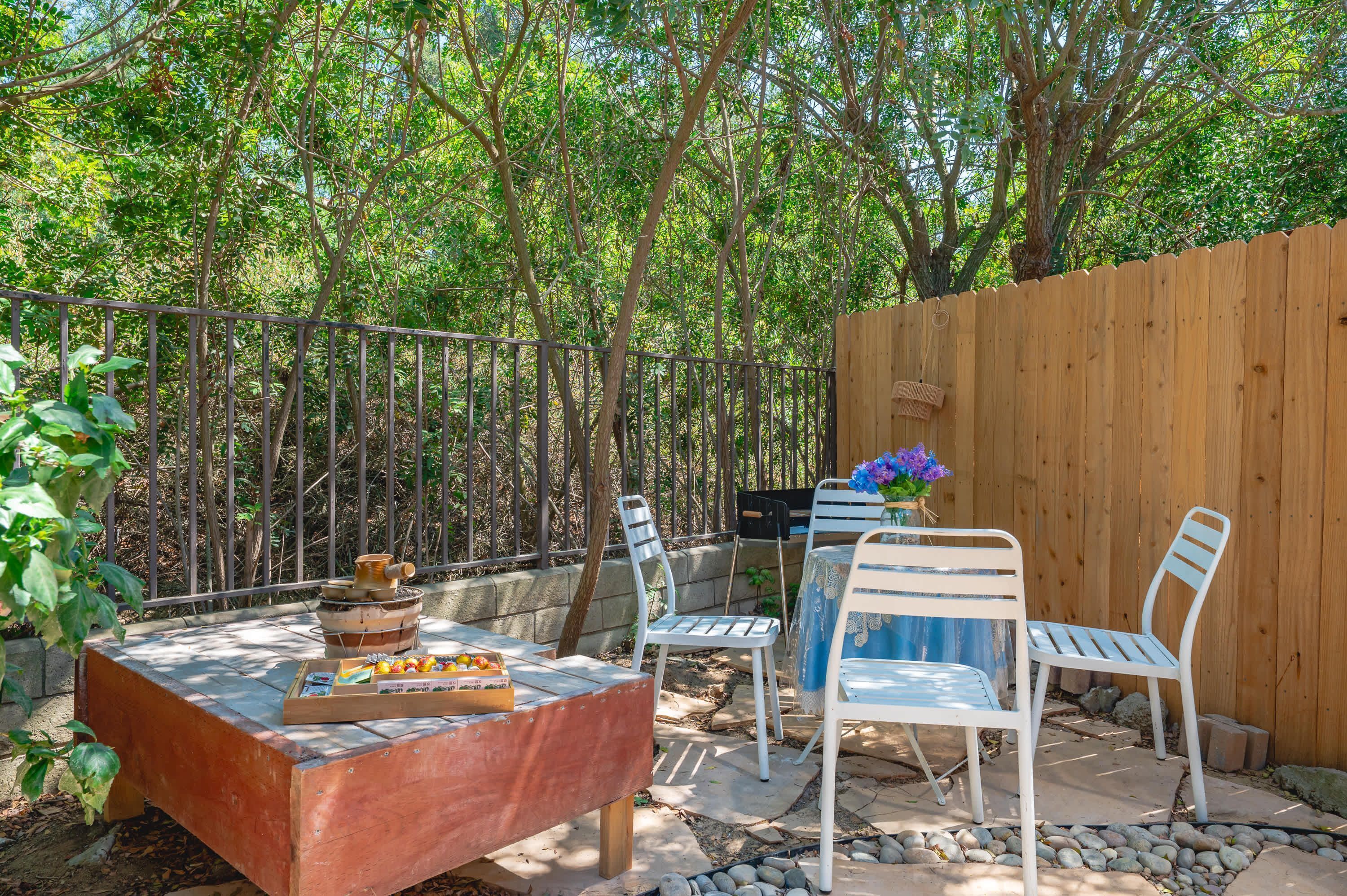 An outdoor patio area with a wooden table, two white chairs, a small grill, and a wooden fence.