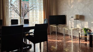 Ein Hotelzimmer mit Esstisch, Stühlen, Fernseher, Lampe und einer Vase mit Zweigen.