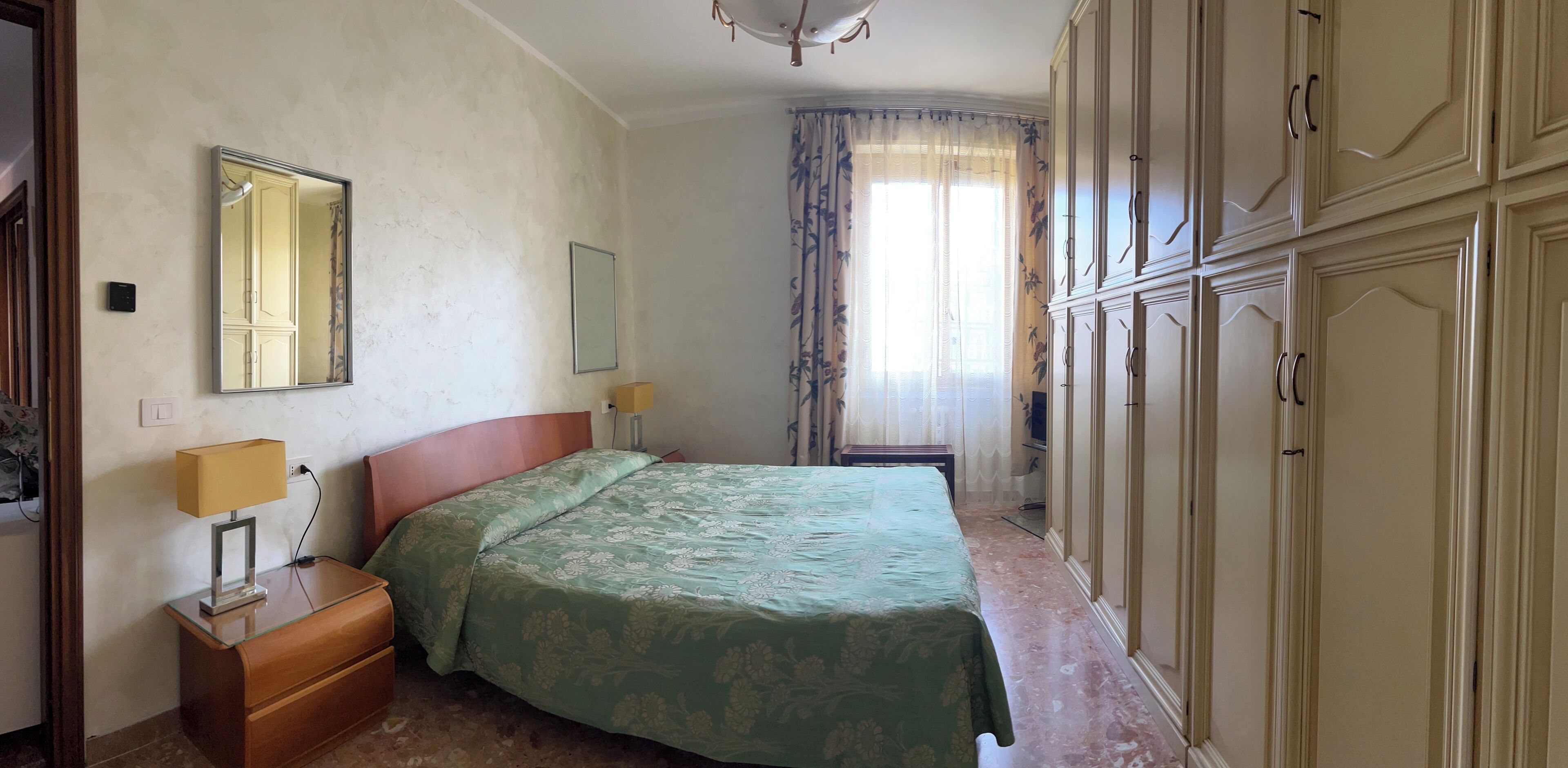 A bedroom with a bed, bedside lamps, a mirror, a wardrobe, and a window with curtains.