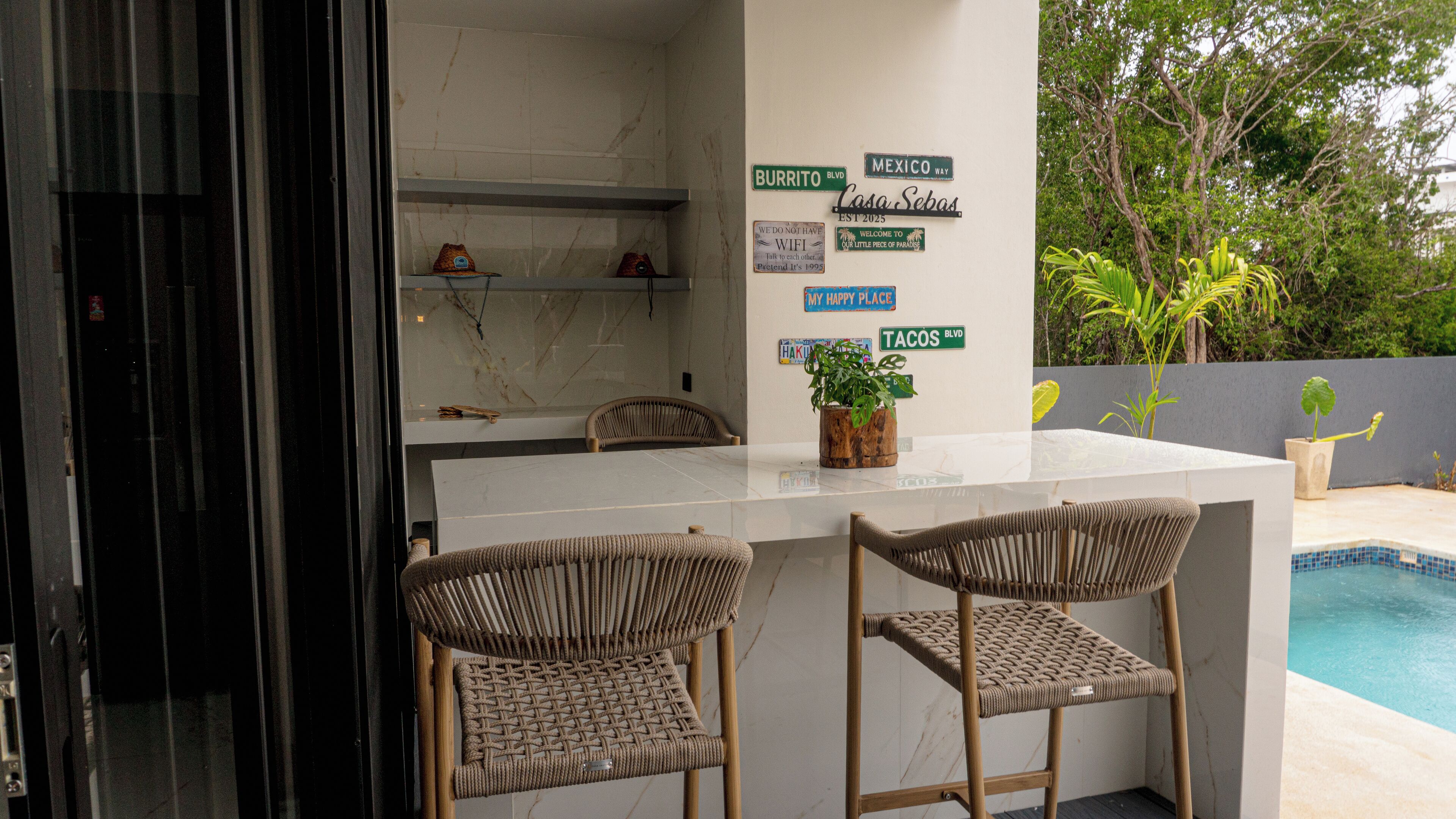 A modern outdoor bar area with two wicker chairs, a marble countertop, and a potted plant.