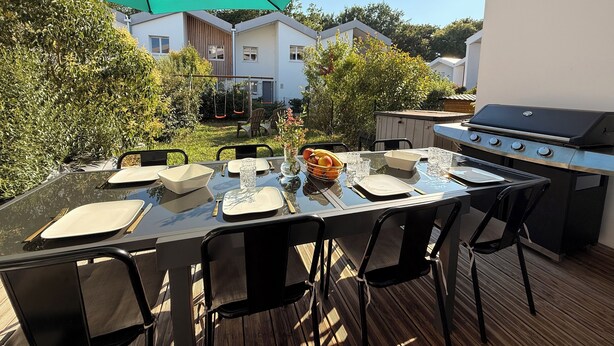 Un área de comedor al aire libre con una mesa lista para cuatro personas, rodeada de sillas, bajo un paraguas verde. Se ve una parrilla de barbacoa a la derecha.