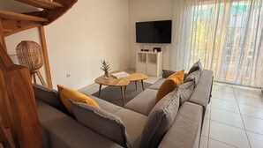 Una sala de estar moderna con un sofá gris, una escalera de madera, un televisor de pantalla plana y un ventanal con cortinas translúcidas.
