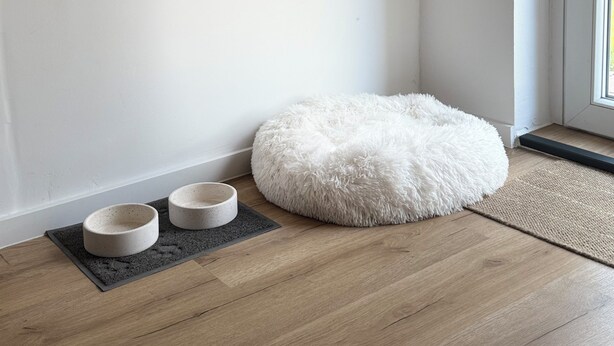 A corner of a room with a wooden floor, a white fluffy pet bed, and two pet bowls on a mat. - Cozy vacation apartment in the heath (Hodenhagen)