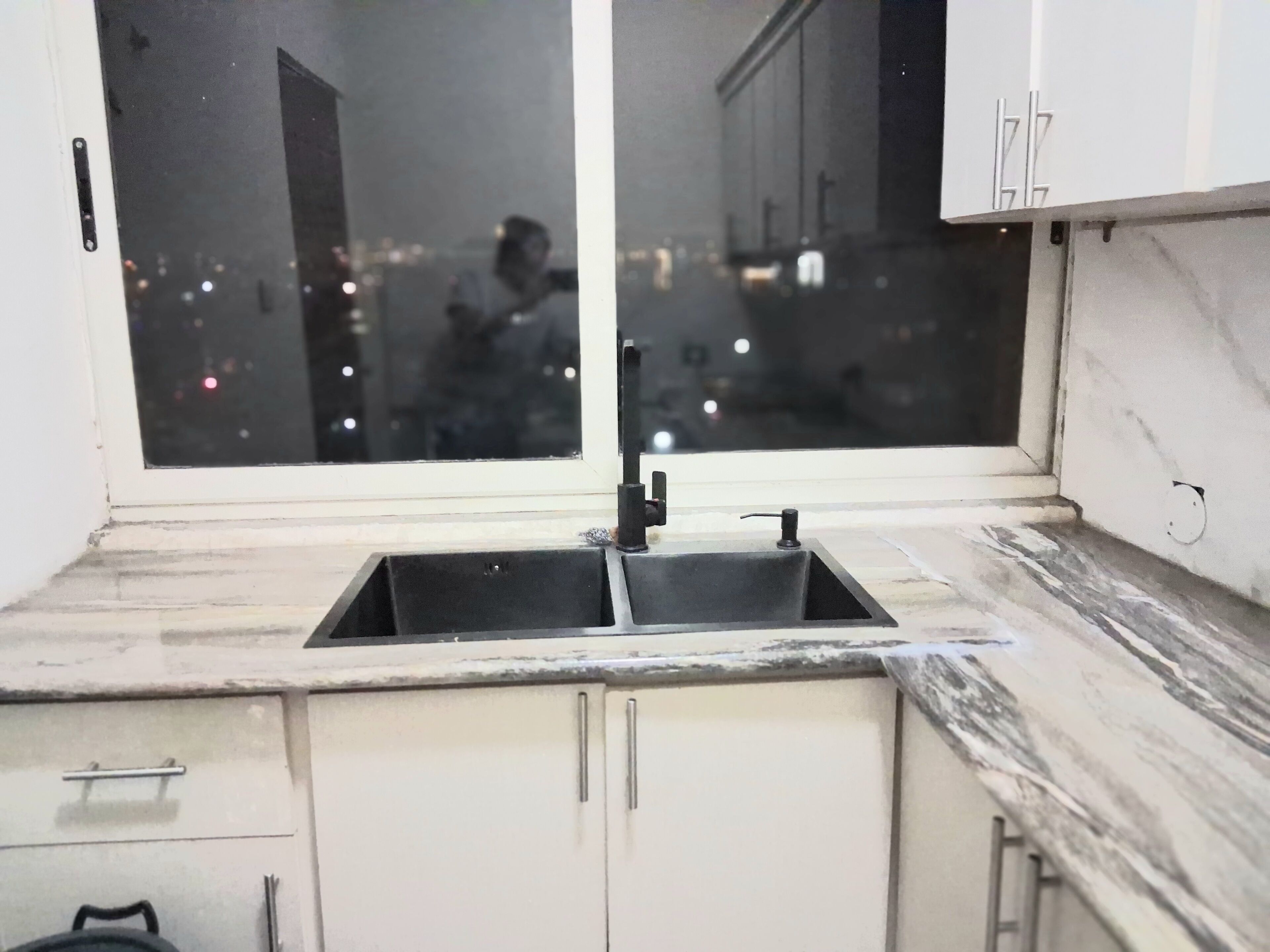 Une cuisine dotée d’un évier à deux bacs, de meubles blancs et d’une fenêtre donnant sur une vue nocturne de la ville.