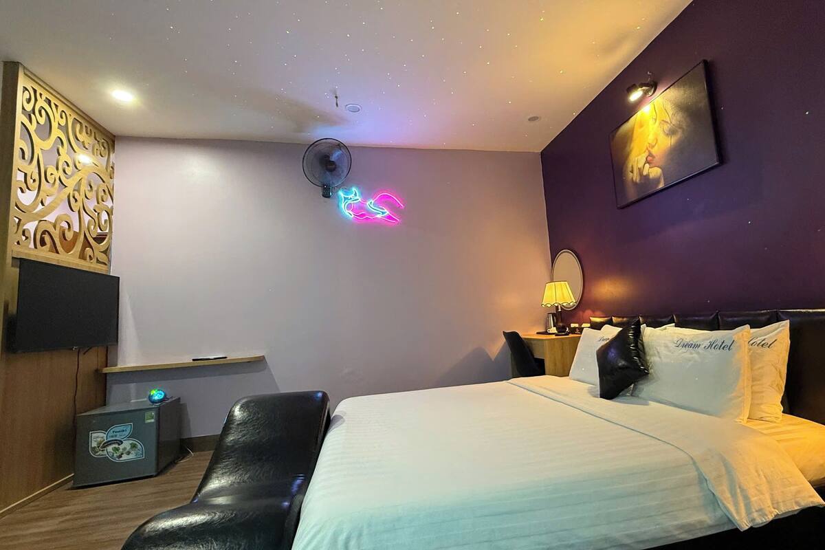 Una habitación de hotel con una cama, un escritorio, una silla, un ventilador de techo, un televisor de pared y un letrero de neón.