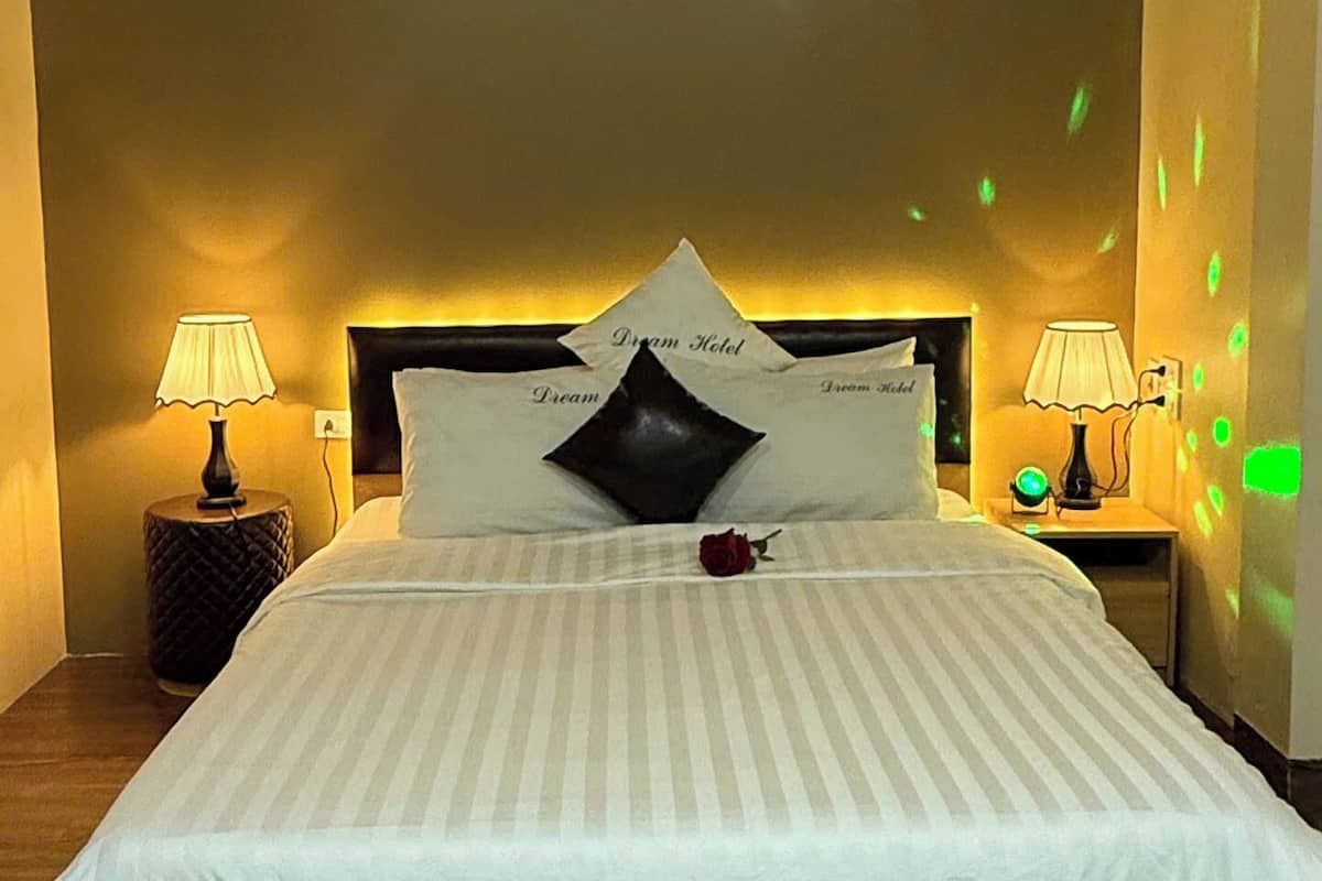 Habitación de hotel con una cama grande, dos mesitas de noche con lámparas, un cabecero con el nombre del hotel y una sola rosa decorativa sobre la cama.