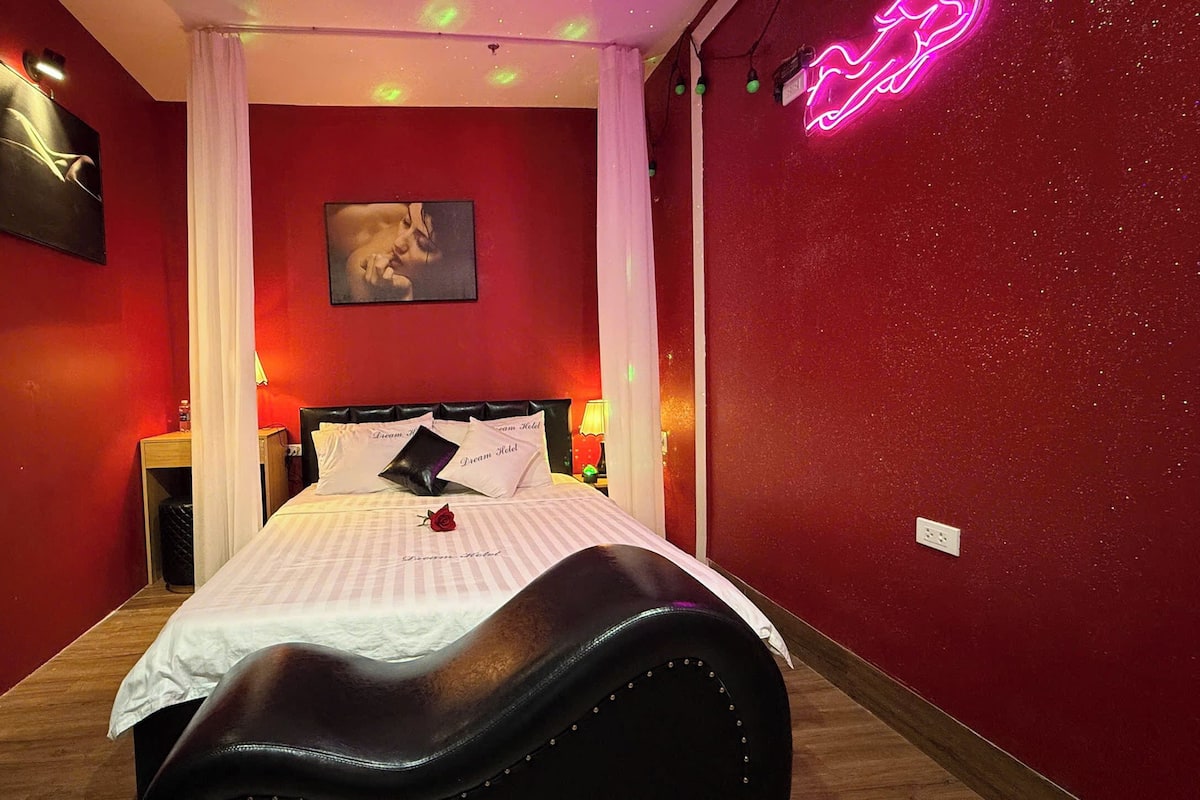 Habitación de hotel con pared roja, una cama con ropa de cama blanca, un sillón de cuero negro, un letrero de neón y un cuadro en la pared.