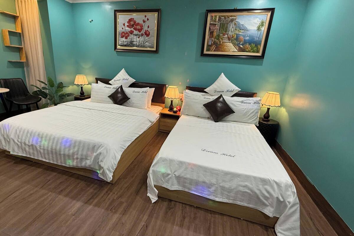 Habitación de hotel con dos camas, cada una con un cojín que dice "Dream Hotel", una mesita con una silla, una lámpara y un cuadro en la pared.