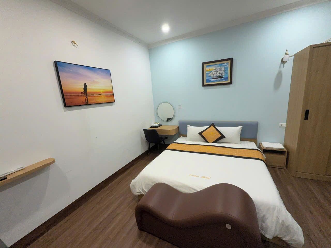 A hotel room with a bed, a desk, a chair, a wardrobe, and two paintings on the walls.