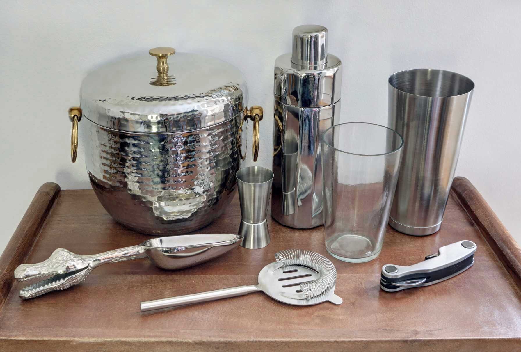 A tray with a large silver cocktail shaker, a smaller shaker, a glass, a spoon, a strainer, and a bottle opener.