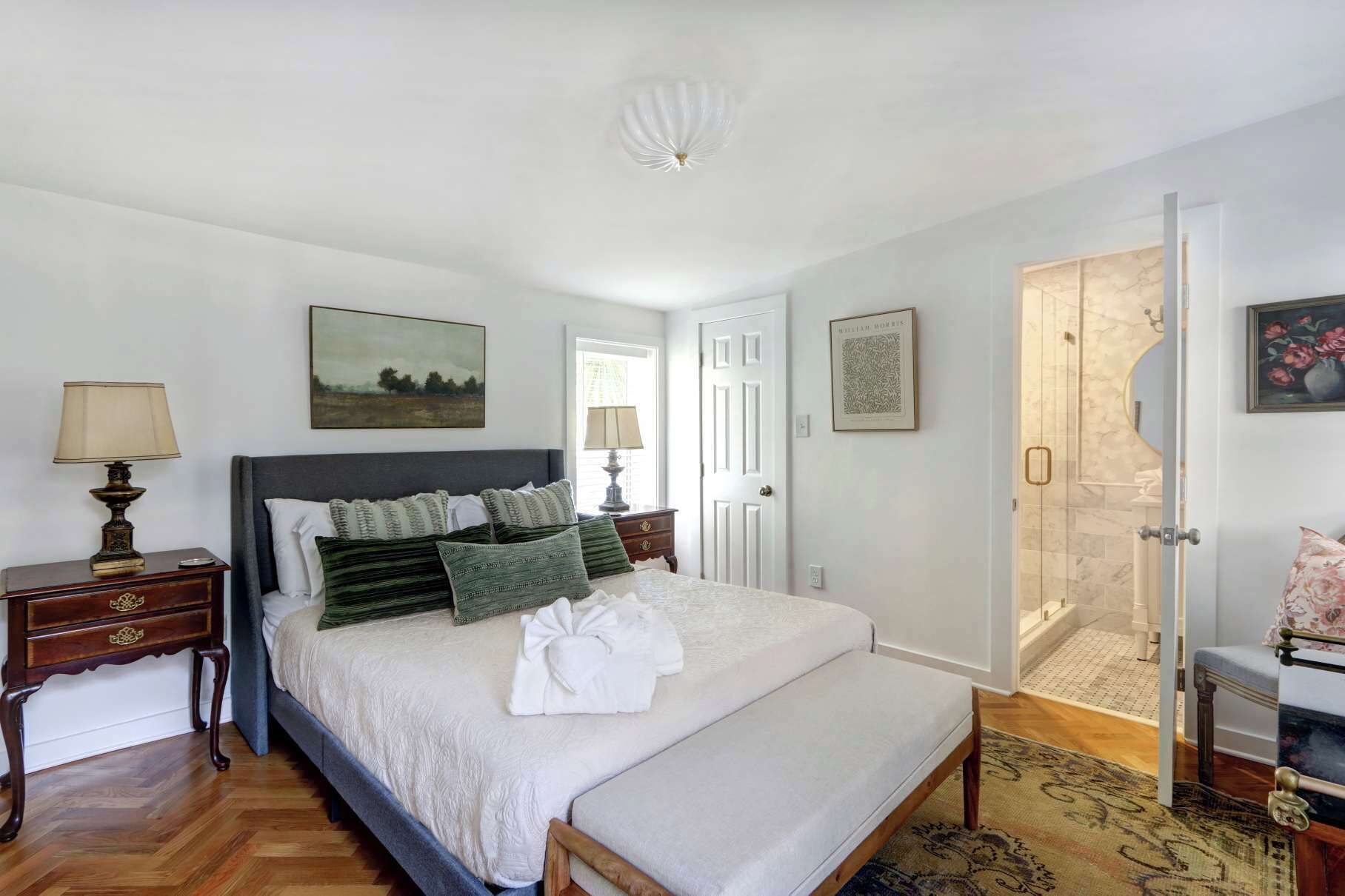 A bedroom with a large bed, a bedside table with a lamp, a door leading to a bathroom, and a painting on the wall.