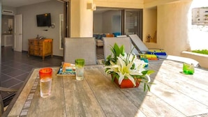 Une terrasse ensoleillée avec une table en bois, une plante en pot et une vue sur l’intérieur.