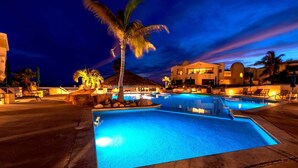 A nighttime swimming pool area with multiple pools, palm trees, and resort buildings.