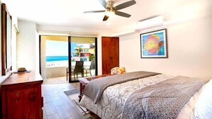 Une chambre avec un lit, un ventilateur de plafond, une armoire en bois, un tableau encadré et une vue sur la plage, visible à travers une porte coulissante.