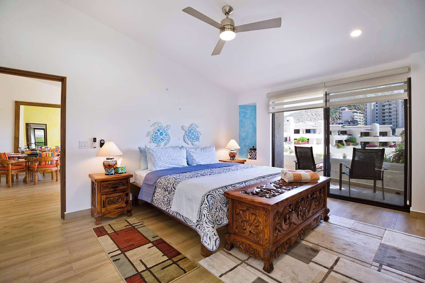 A bedroom with a bed, two bedside tables, a ceiling fan, a large window, and a view of the outside.