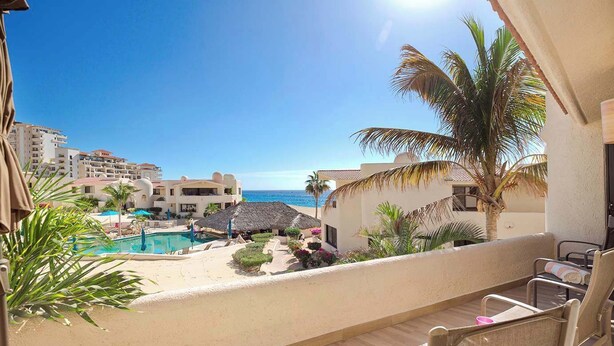 Un balcon avec vue sur une piscine et une plage, entouré de palmiers et surplombant un ciel bleu azur.