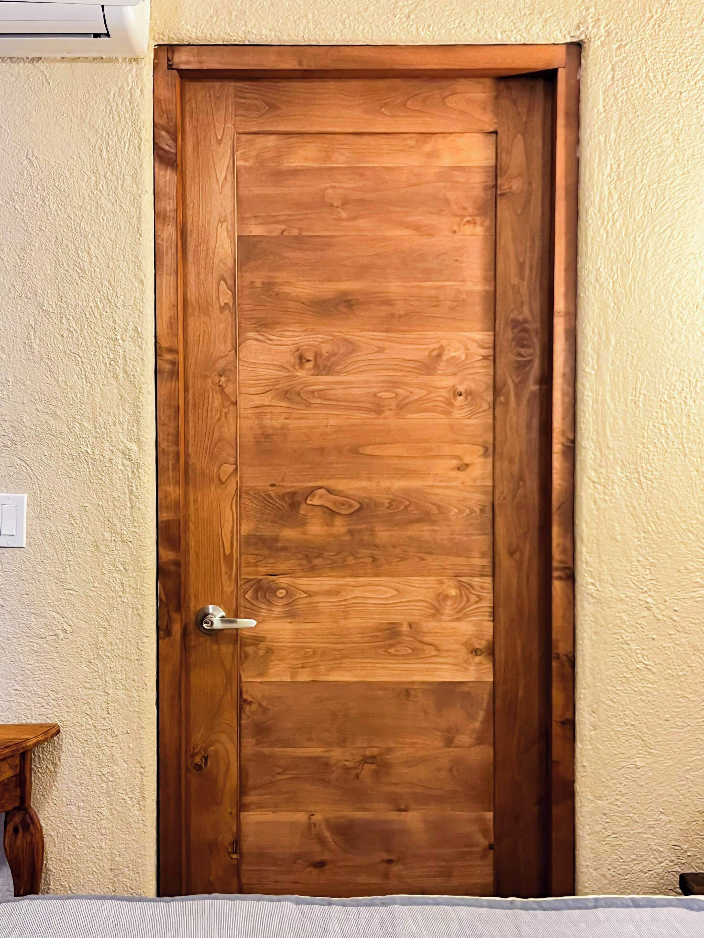 Une porte en bois avec une poignée argentée, située dans une pièce aux murs texturés et dont l’interrupteur est visible.