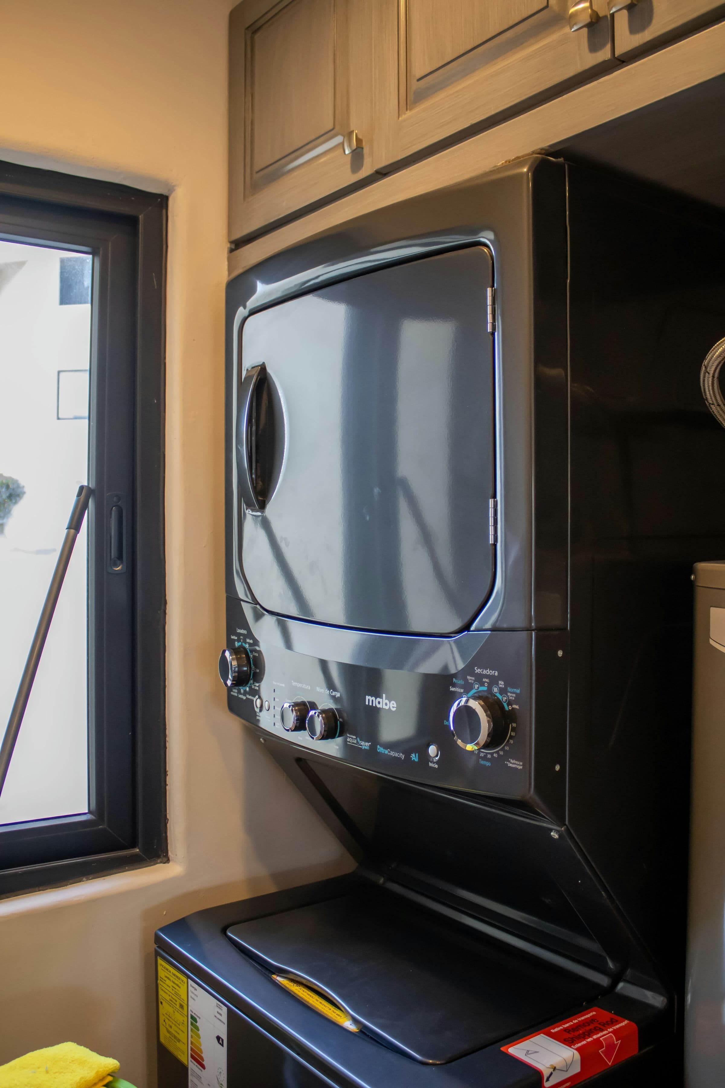 Lavadora compacta y moderna con mandos y pantalla digital, ubicada en un lavadero pequeño con armarios de madera y una ventana.