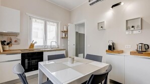Une cuisine moderne avec des meubles blancs, une table à manger et des chaises. On y trouve une fenêtre avec des rideaux, un four encastré et une machine à café sur le plan de travail.